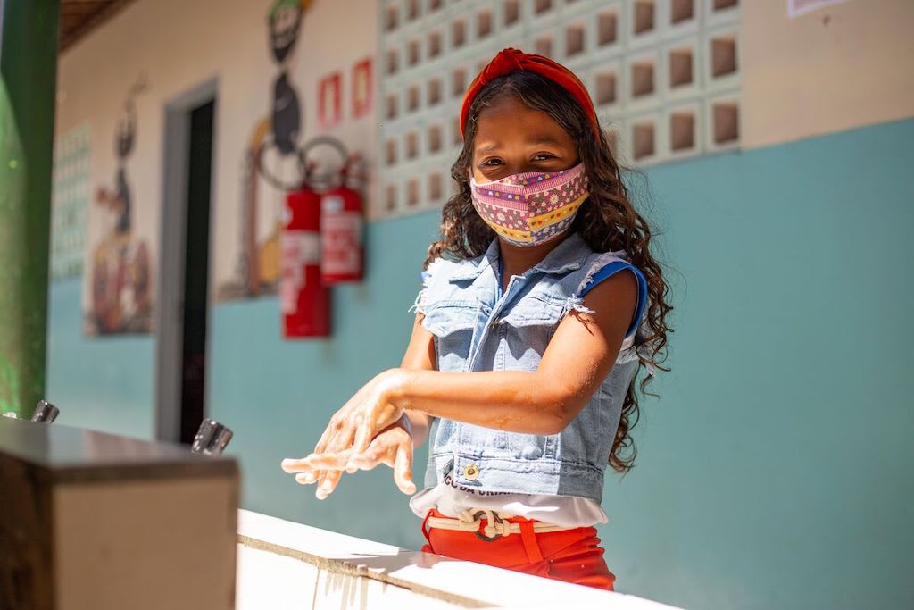 A girl in a denim top washes her hands while wearing a protective face mask.